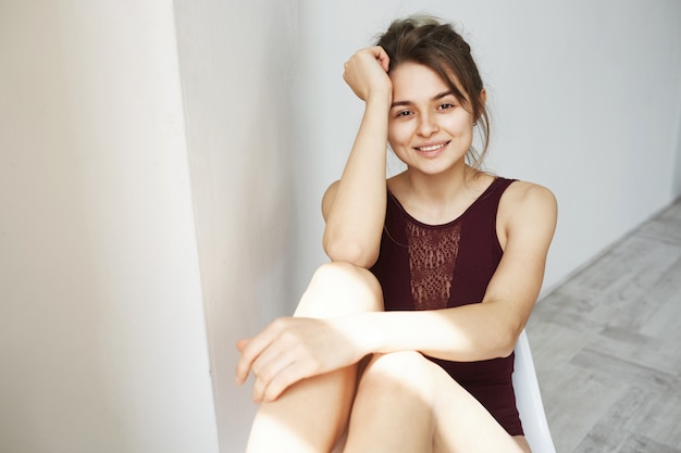 Portrait de la belle jeune fille tendre en maillot de bain vineux souriant posant sur une chaise sur un mur blanc.