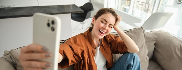 Photo gratuite portrait d'une belle jeune mannequin heureuse assise sur le canapé avec un smartphone en train de se faire un selfie