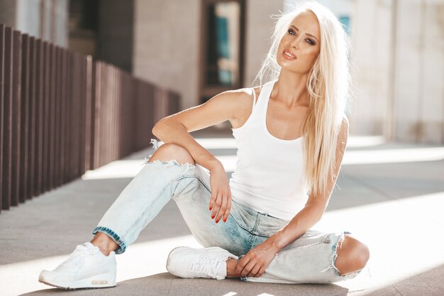 Portrait de la belle jolie fille blonde en T-shirt blanc et jeans posant à l'extérieur. Jolie fille assise sur l'asphalte dans la rue