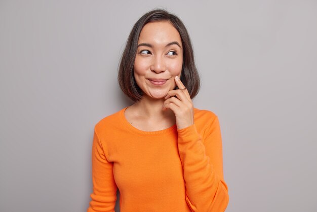 Portrait d'une belle mannequin asiatique se souvient de son premier rendez-vous avec son mari a une expression rêveuse et heureuse, des sourires doucement concentrés portent des poses de pull orange décontractées à l'intérieur contre un mur gris