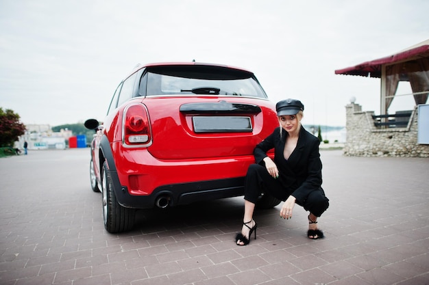 Portrait d'une belle mannequin blonde sexy en casquette et en tout noir avec un maquillage lumineux près d'une voiture de ville rouge