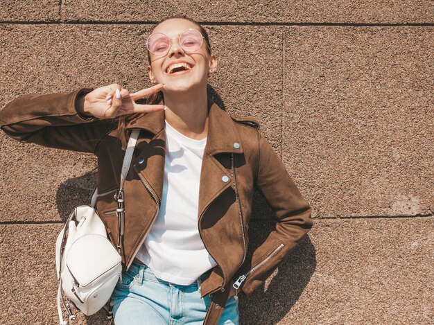 Portrait de la belle mannequin brune souriante habillée en veste d'été hipster et vêtements jeans près du mur Femme drôle et positive montrant le signe de la paix