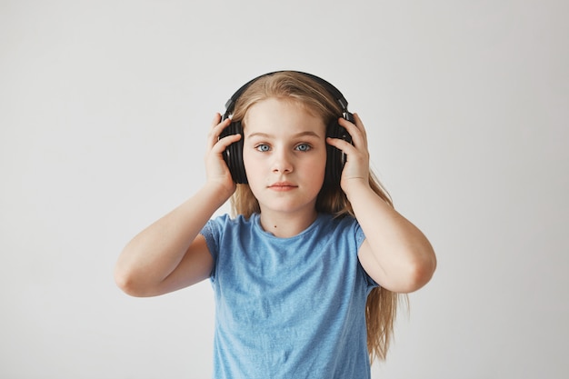 Portrait de la belle petite fille blonde aux cheveux longs et aux yeux bleus portant de gros écouteurs, le tenant avec les mains, écoutant de la musique avec une expression détendue.