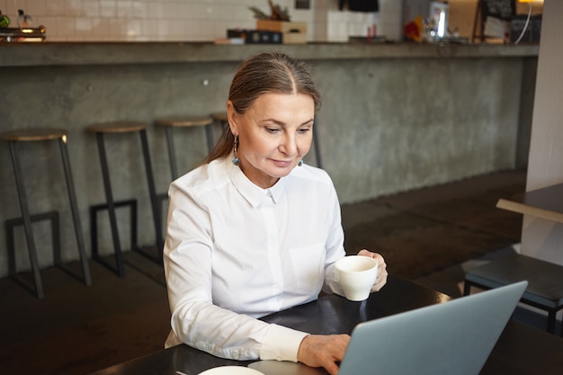 Portrait de bon à la recherche d'une élégante pigiste de 60 ans en chemise blanche ayant une pause à la cafétéria, assis à table devant un ordinateur portable ouvert, boire du thé et surfer sur Internet