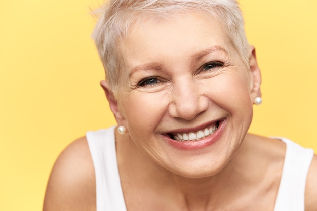 Portrait de bonne à la femme européenne d'âge moyen gaie avec une coiffure élégante portant un réservoir blanc, exprimant des émotions positives, souriant largement, montrant des dents droites, heureux de recevoir de bonnes nouvelles