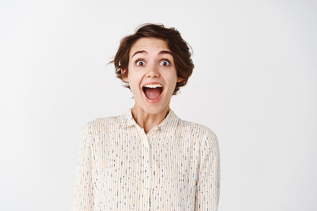 Portrait candide d'une jeune fille naturelle criant de joie et d'étonnement, l'air surpris de découvrir l'offre promotionnelle, debout sur un mur blanc