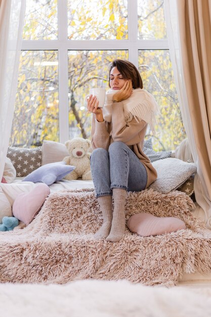 Portrait chaleureux de femme assise sur le rebord de la fenêtre avec une tasse de café thé chaud portant chandail et foulard blanc