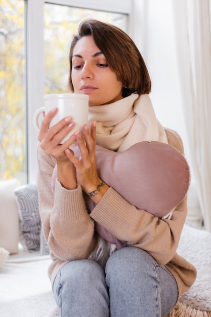 Portrait chaleureux de femme assise sur le rebord de la fenêtre avec une tasse de café thé chaud portant chandail et foulard blanc