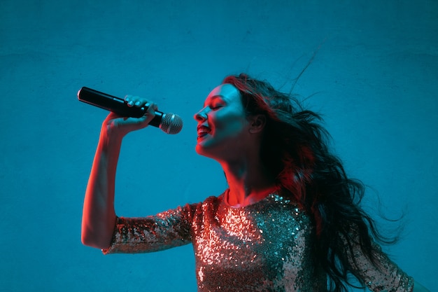 Portrait de chanteuse caucasienne isolé sur fond bleu studio en néon. Beau modèle féminin en robe lumineuse avec microphone. Concept d'émotions humaines, expression faciale, publicité, musique, art.