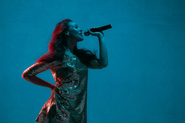 Portrait de chanteuse caucasienne isolé sur fond bleu studio en néon. Beau modèle féminin en robe lumineuse avec microphone. Concept d'émotions humaines, expression faciale, publicité, musique, art.