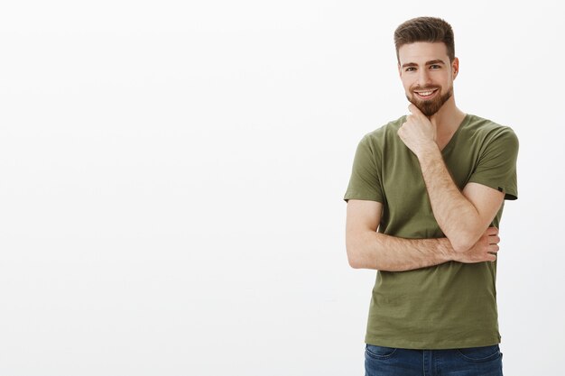 Portrait de charmant charismasitc homme barbu caucasien aux yeux bleus se frottant le menton comme souriant ravi posant à droite de l'espace de copie sur un mur blanc, entendant une excellente idée