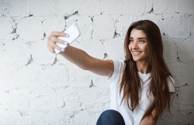 Portrait de charmant modèle féminin européen, faisant selfie sur smartphone près du mur de briques blanches, souriant joyeusement. La blogueuse à la mode prend une photo pour la publier dans son blog. Elle a beaucoup de fans.