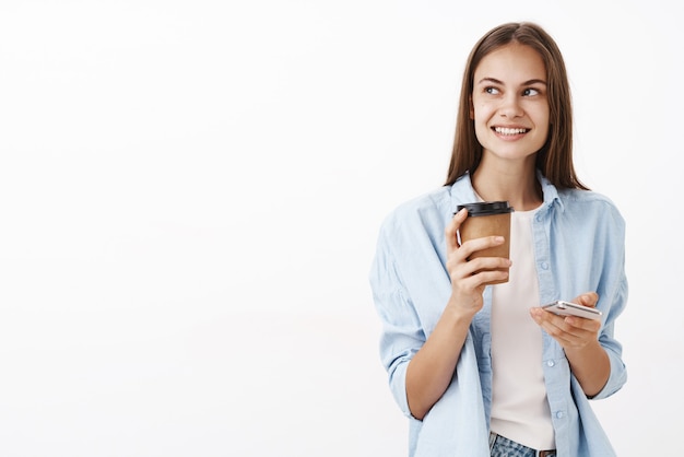 Portrait De Charmante Employée Intelligente Et élégante Tenant Une Tasse De Papier De Boisson Et Smartphone Regardant à Gauche Avec Un Sourire Heureux D'avoir Plan