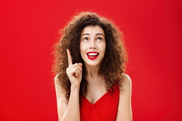 Portrait d'une charmante femme idiote ravie avec une coiffure frisée en robe levant l'index dans un geste eurêka souriant joyeusement en levant un bon plan ou une idée posant charismatique sur un mur rouge.