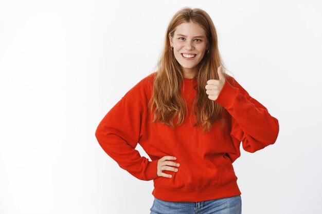 Portrait d'une charmante femme mignonne sincère en pull rouge chaud montrant le geste du pouce tenant la main sur la taille et souriante donnant une réponse positive, aimant l'idée