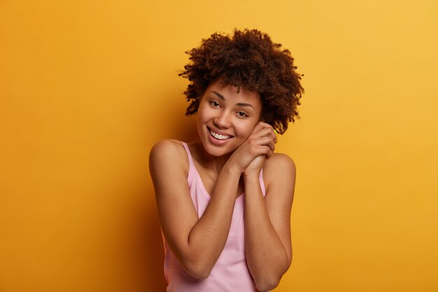 Portrait de charmante femme à la recherche agréable incline la tête et regarde positivement, a une expression sincère et heureuse, un regard confiant, pose contre le mur jaune, porte une chemise décontractée