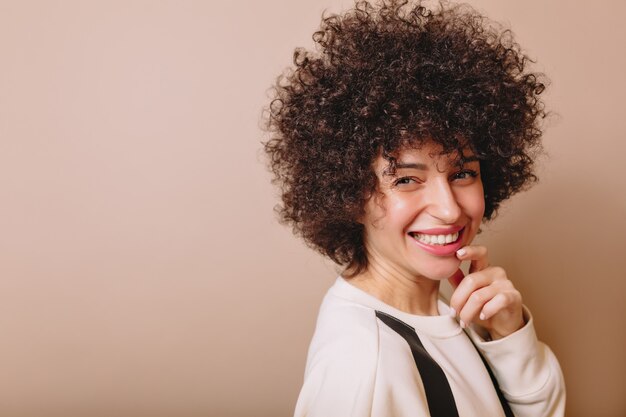 Portrait de charmante femme riant avec des boucles et un sourire merveilleux pose sur beige et tient une main près du visage