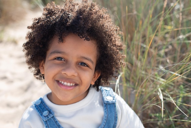 Portrait De Charmante Fille Afro-américaine Sur La Plage. Modèle Féminin Aux Cheveux Bouclés En Jeans Assis Sur Le Sable, Regardant La Caméra. Portrait, Notion De Beauté