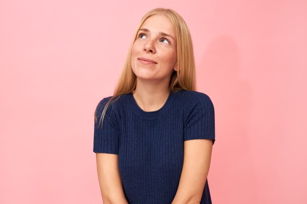 Portrait de charmante jeune femme aux longs cheveux blonds posant isolé habillé en t-shirt bleu foncé regardant avec un sourire rêveur, pensant à l'avenir