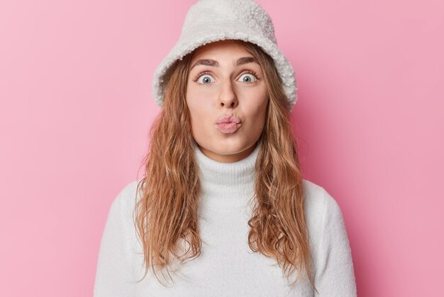 Portrait d'une charmante jeune femme impressionnée garde les lèvres pliées, les coups s'embrassent à la caméra a un look attrayant, les cheveux longs portent un panama de fourrure et un col roulé isolés sur fond rose. Expressions du visage humain