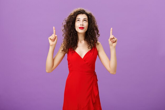 Portrait d'une charmante jeune femme intriguée et curieuse avec une coiffure frisée en robe de soirée rouge élégante regardant et pointant vers le haut souriante intéressée et ravie posant sur fond violet.