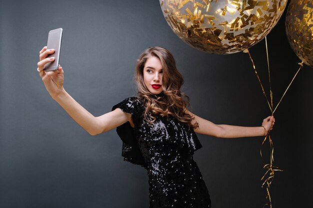 Portrait charmante jeune femme en robe de luxe noire, avec de longs cheveux bruns bouclés, des lèvres rouges prenant selfie avec de gros ballons pleins de guirlandes dorées. Magnifique modèle, célébration.