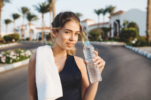 Portrait charmante jeune femme séduisante en tenue de sport avec une bouteille d'eau à la formation en matinée ensoleillée sur la rue avec des palmiers dans la ville tropicale. Outwork, style de vie sportif, bonne humeur.
