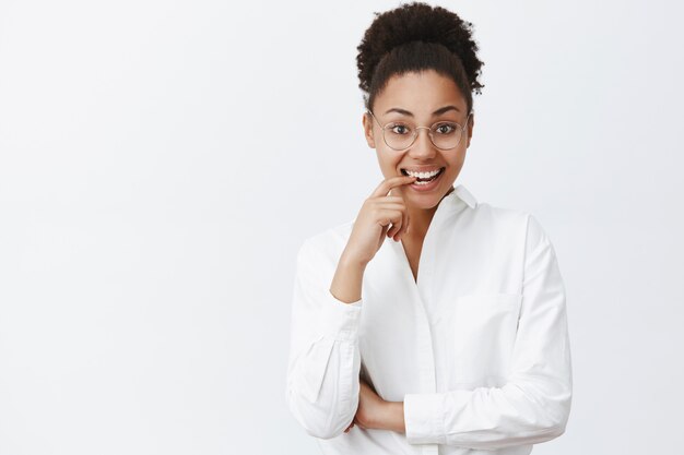 Portrait de charmante tutrice à la peau foncée émotive et féminine en chemise à col blanc et lunettes, mordant le doigt par curiosité et souriant largement, regardant intriguée et intéressée