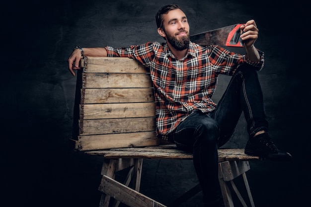 Portrait de charpentier barbu s'habille d'une chemise à carreaux et d'un jean tient une scie à main sur une épaule.