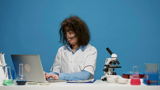 Portrait d'une chimiste folle travaillant sur un ordinateur portable au bureau, étant drôle et maladroite en studio. Scientifique fou fou utilisant un ordinateur et faisant des expressions faciales stupides et stupides, ayant les cheveux en désordre.