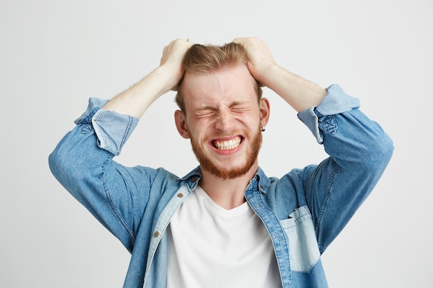 Portrait de colère jeune homme en colère touchant ses cheveux serré les dents sur fond blanc. Yeux fermés.