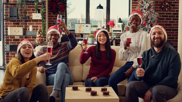 Portrait de collègues festifs trinquant avec de l'alcool pour célébrer les vacances de noël avec des toasts ou des acclamations. Les gens célèbrent l'événement de fête d'hiver avec une boisson au vin dans un bureau décoré.