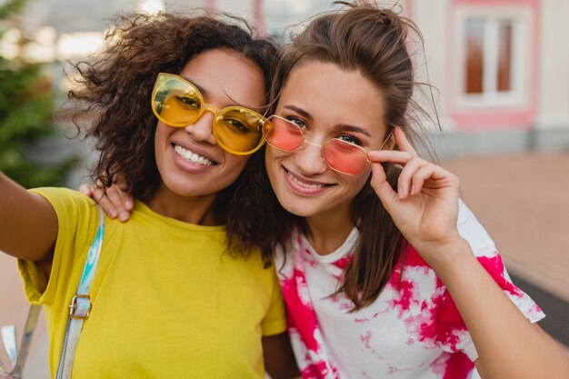 Portrait coloré d'amis heureux jeunes filles souriant assis dans la rue en prenant selfie photo sur téléphone mobile, les femmes s'amusant ensemble