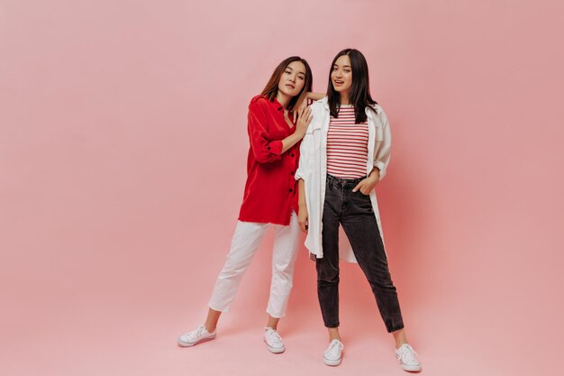 Portrait complet de deux femmes asiatiques sur fond rose Fille aux cheveux courts en pantalon blanc, chemise rouge et dame brune en jeans noirs posent sur isolé