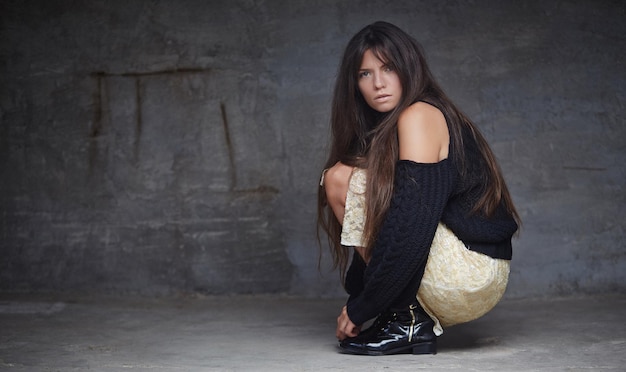 Photo gratuite portrait complet du corps d'une femme de mode brune assise sur le sol sur un mur gris.