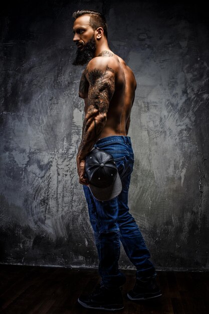 Portrait complet du corps d'un homme barbu tatoué musclé en jeans bleus sur un mur gris.