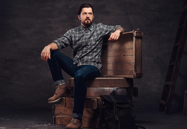 Portrait complet du corps d'un homme hipster tatoué d'âge moyen vêtu d'une chemise à carreaux et d'un jean, assis sur des caisses en bois dans un studio. Isolé sur un fond texturé sombre.