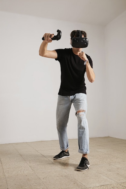 Portrait complet du corps d'un jeune joueur vêtu de lunettes et de jeans VR et d'un t-shirt noir sans étiquette jouant à un jeu dans une pièce aux murs blancs et au parquet clair.