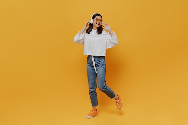 Portrait complet d'une femme brune en pantalon en jean à capuche blanc et lunettes de soleil orange chantant et écoutant de la musique dans des écouteurs sur fond orange