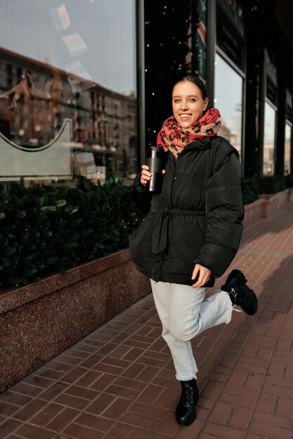 Portrait complet d'une fille heureuse et excitée portant un jean, une veste chaude foncée et une écharpe rouge marche dans une rue ensoleillée de la ville avec du café