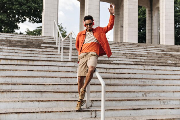 Portrait complet d'un homme brun bouclé à la peau foncée en short beige et veste orange posant et s'amusant dans les escaliers à l'extérieur Jeune homme à lunettes de soleil écoute de la musique dans les écouteurs