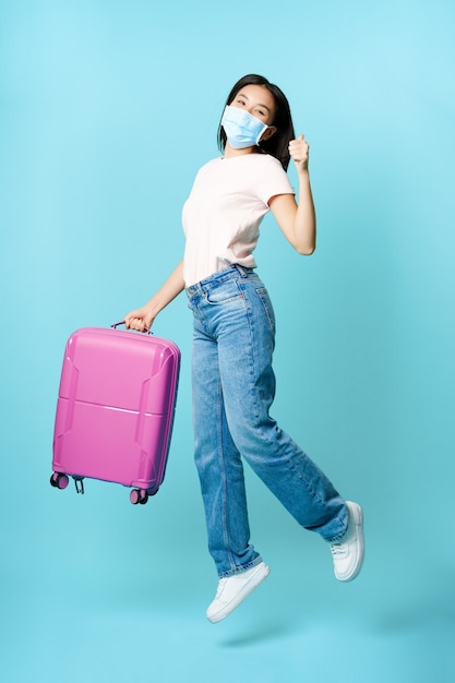 Portrait complet d'une touriste asiatique heureuse, sautant avec une valise, montrant les pouces vers le haut, profitant de vacances en tournée, fond bleu.