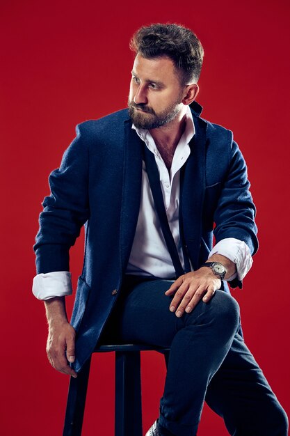 Portrait de concept de beauté masculine d'un jeune homme à la mode avec une coupe de cheveux élégante portant un costume à la mode posant sur un mur rouge