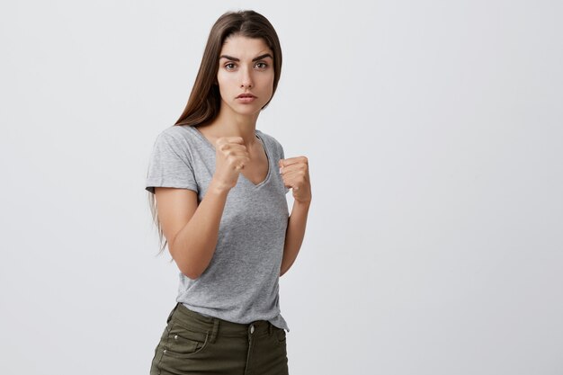 Portrait de confiant beau jeune fille caucasienne aux cheveux longs noirs en t-shirt gris et jeans bruns debout en lutte posent avec une expression faciale sérieuse et moyenne.