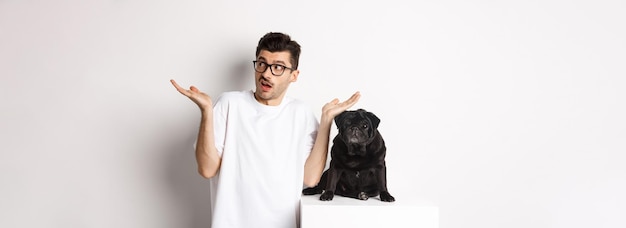 Portrait de confus hipster guy propriétaire de chien haussant les épaules debout près de mignon carlin noir animal blanc backgrou