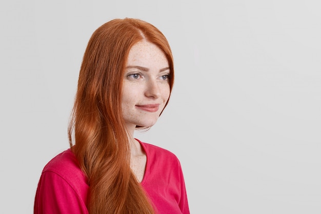 Portrait de côté du modèle féminin de gingembre tacheté réfléchi se souvient de quelques moments agréables de la vie, regarde avec une expression positive dans la distance, isolé sur blanc avec espace de copie