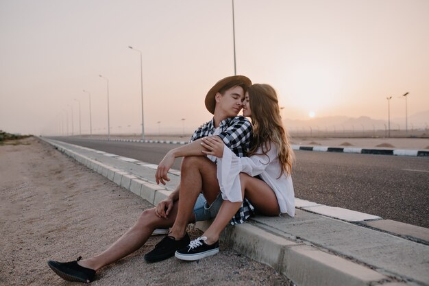 Portrait de couple aimant s'embrasser, assis à côté de l'autoroute après un voyage autour de la ville en week-end d'été. Joyeuse femme aux cheveux longs embrassant doucement son petit ami, se reposant près de la route au coucher du soleil