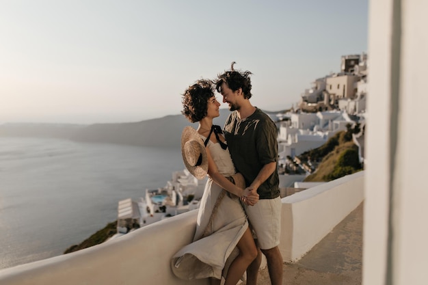 Portrait de couple sur fond de ville grecque Dame brune en robe beige et homme en tenue élégante se regarde avec amour sur fond de mer