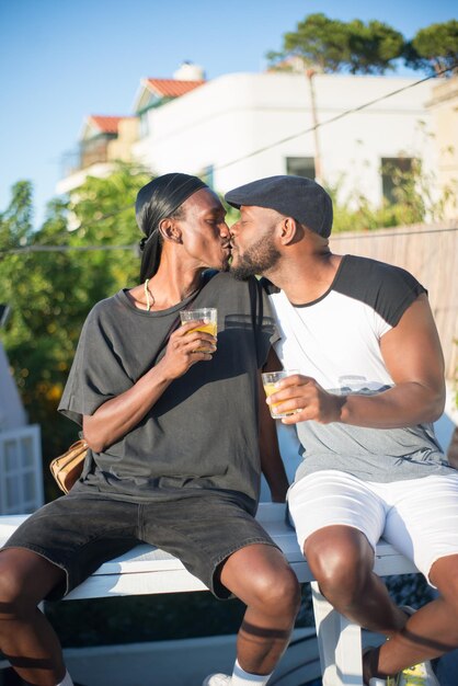 Portrait d'un couple gay africain heureux s'embrassant sur un banc. Deux hommes en tenue décontractée assis sur un banc avec des verres de jus s'embrassant sur les lèvres. L'amour homosexuel et les relations du concept de couples LGBT