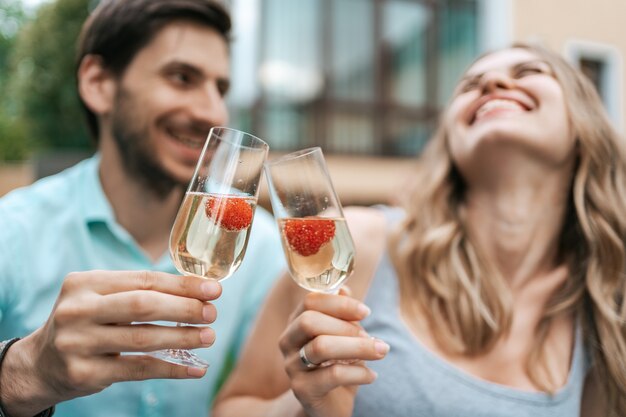 Portrait de couple heureux tinter deux verres avec du vin mousseux et des fraises à l'intérieur avec maison floue sur fond. Célébrer l'amour